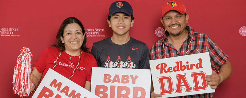Three people holding 'MAMA BIRD,' 'BABY BIRD,' and 'Redbird DAD' signs in front of an Illinois State University backdrop.